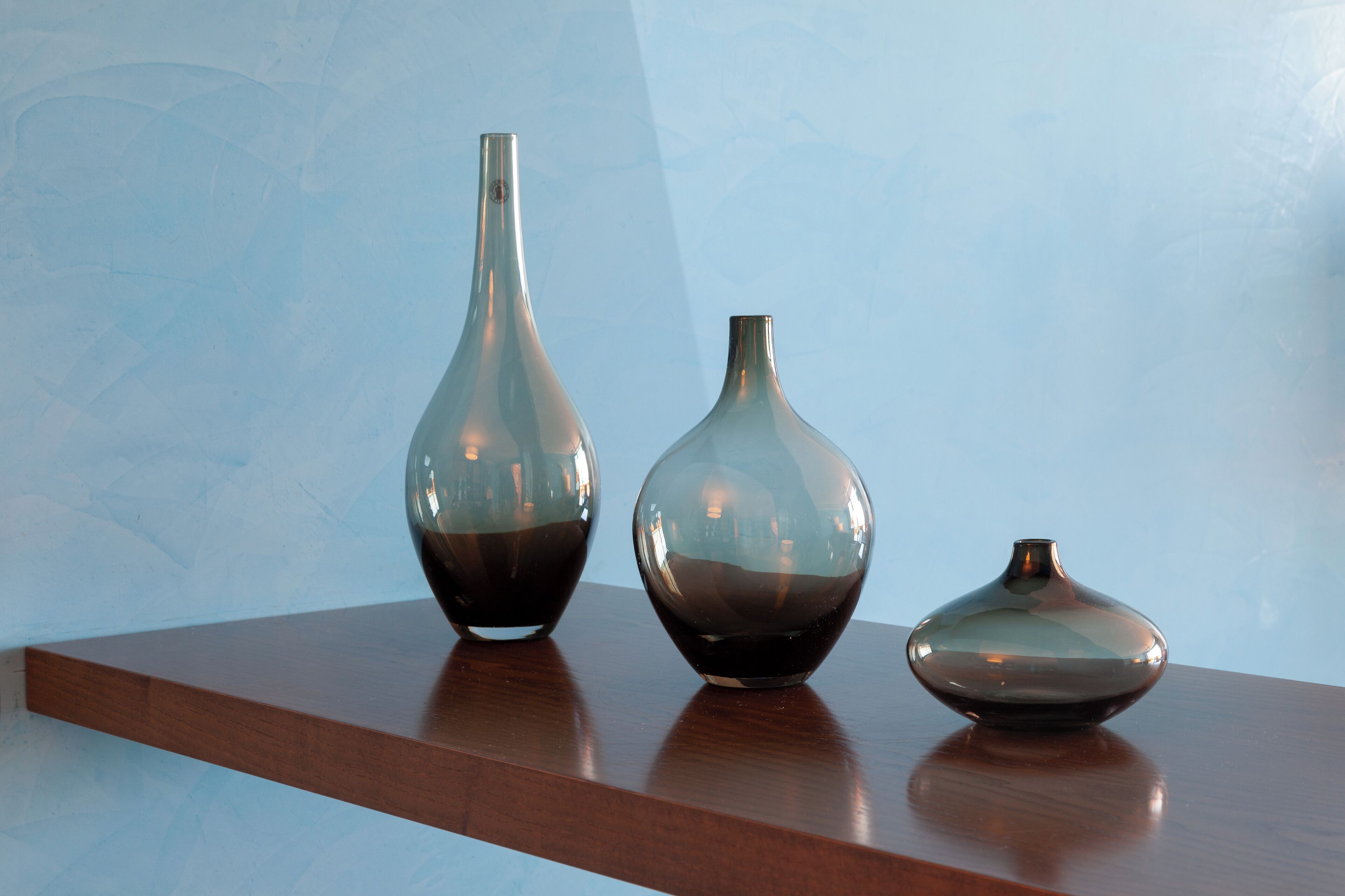 Three glass vases on a wooden shelf against a blue wall.