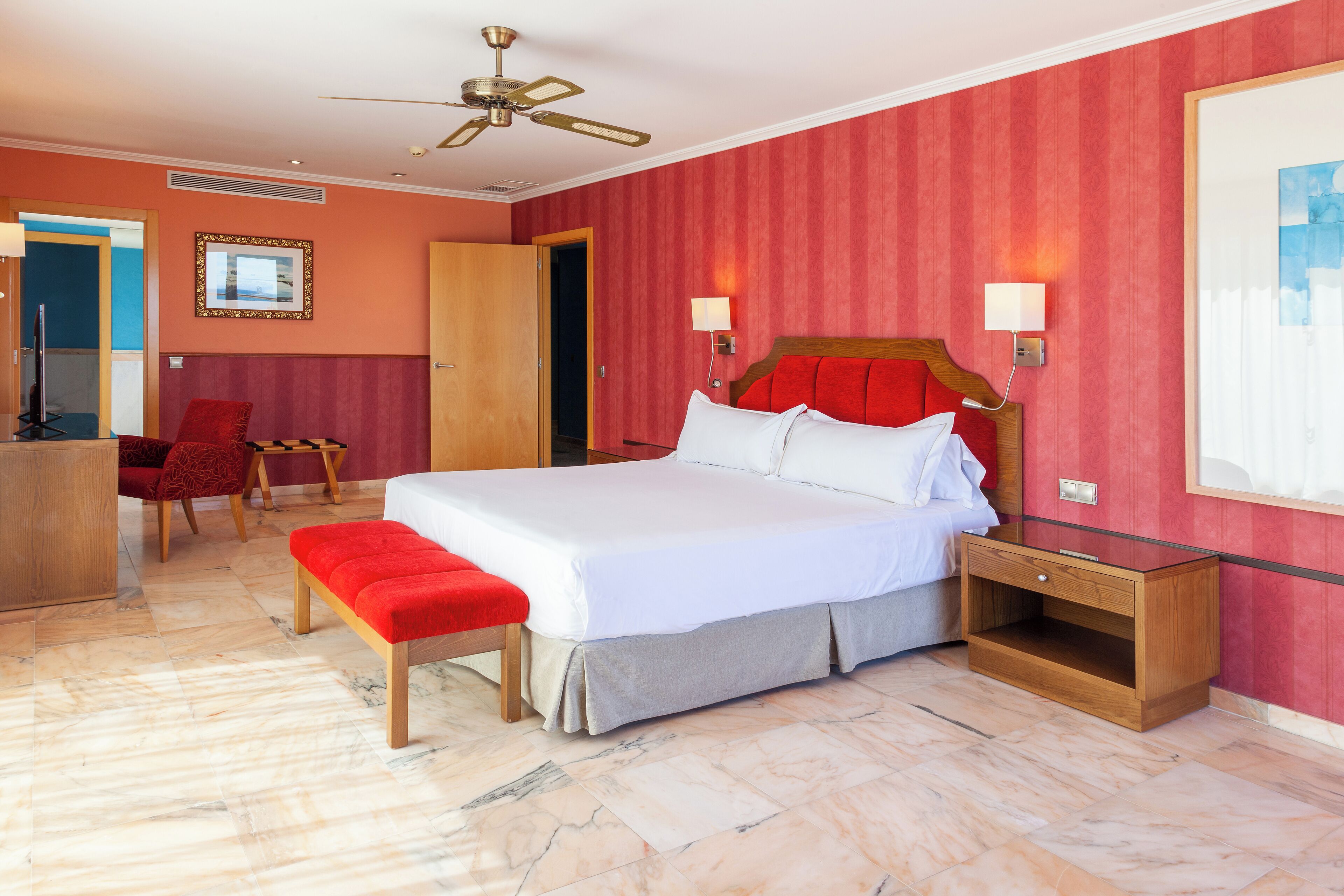 A hotel room with a large bed, a wooden nightstand, a red bench, a desk with a chair, and a ceiling fan.