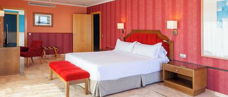 A hotel room with a large bed, a wooden nightstand, a red bench, a desk with a chair, and a ceiling fan.