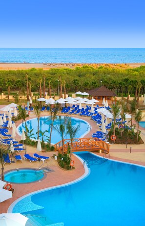 Área de piscina em resort com várias piscinas, espreguiçadeiras, guarda-sóis e palmeiras.