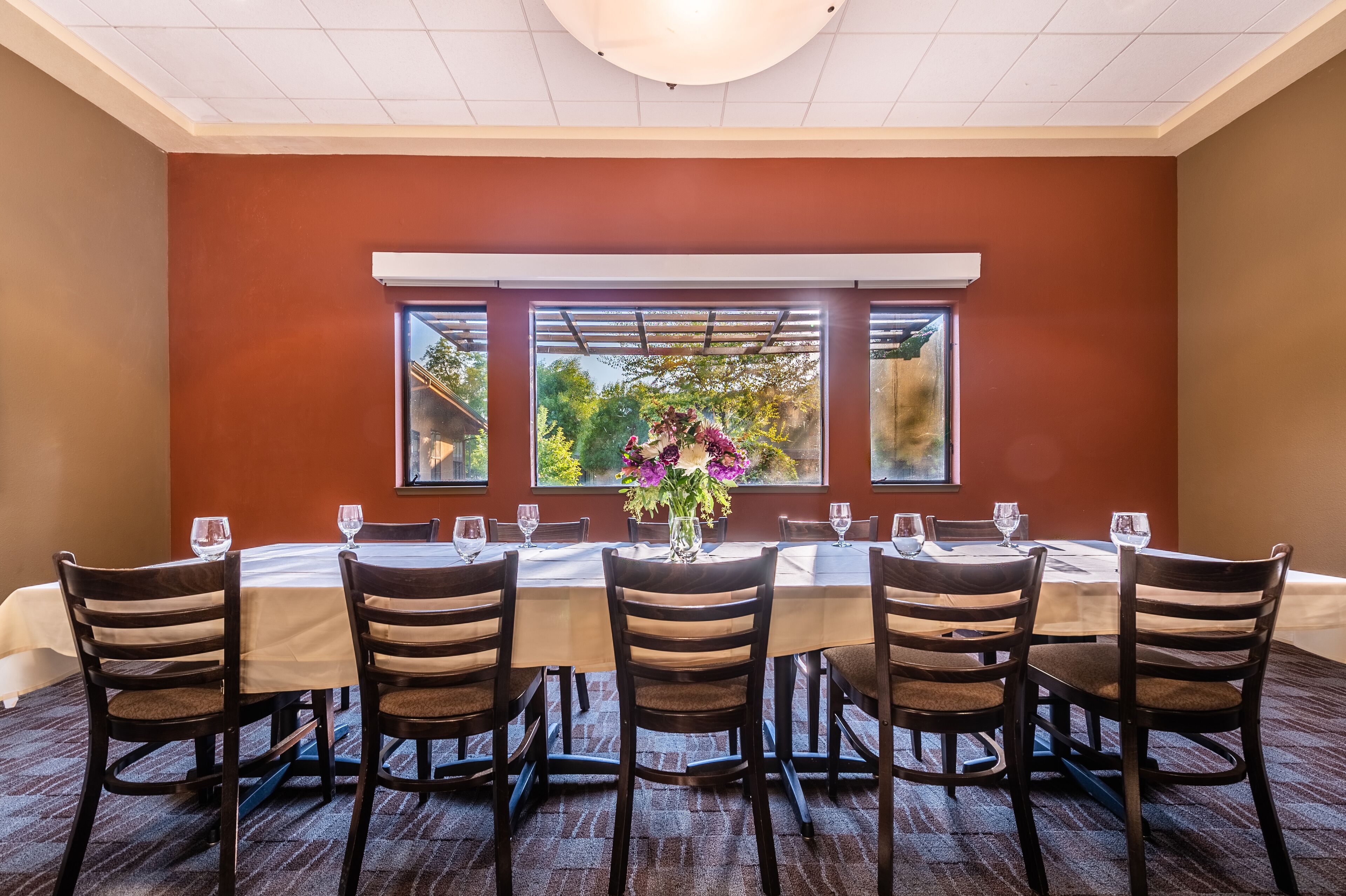A dining area with a long table set for six, surrounded by chairs, with a window view of greenery.