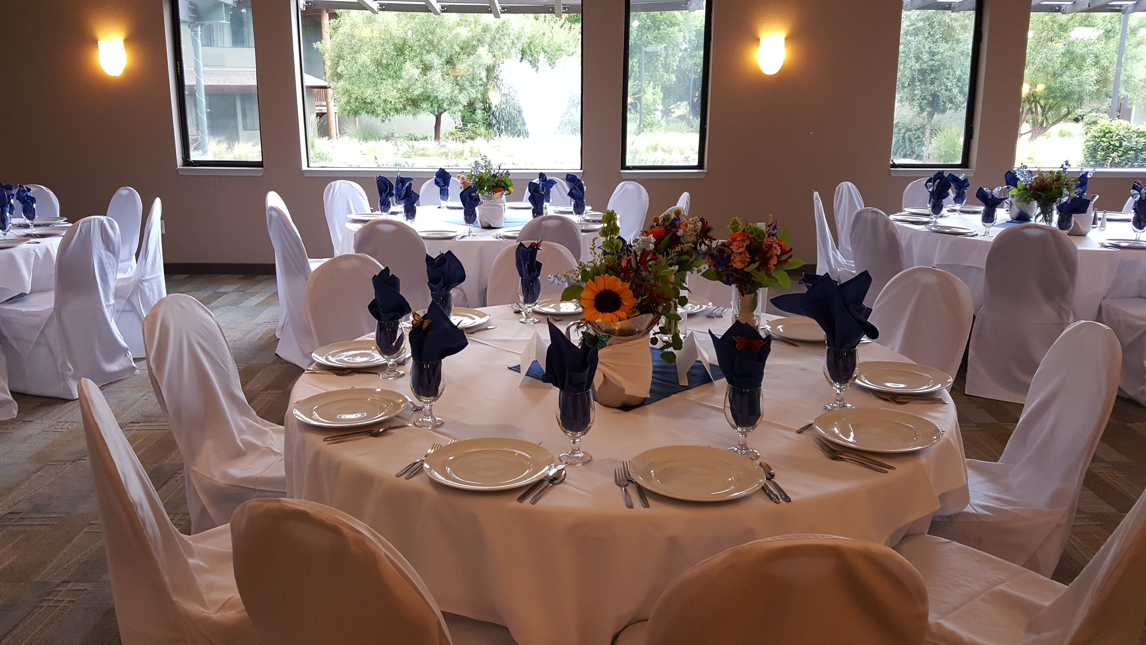 Un salón de banquetes con mesas redondas dispuestas para un evento formal, con manteles blancos, servilletas dobladas con cuidado y centros de mesa florales.