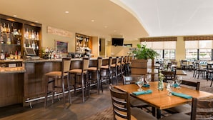A bar area with a wooden bar counter, multiple bar stools, and a variety of bottles and glasses. There are tables and chairs in the dining area with a plant on one table. - Gaia Hotel & Spa Redding, an Ascend Collection Hotel (Anderson)