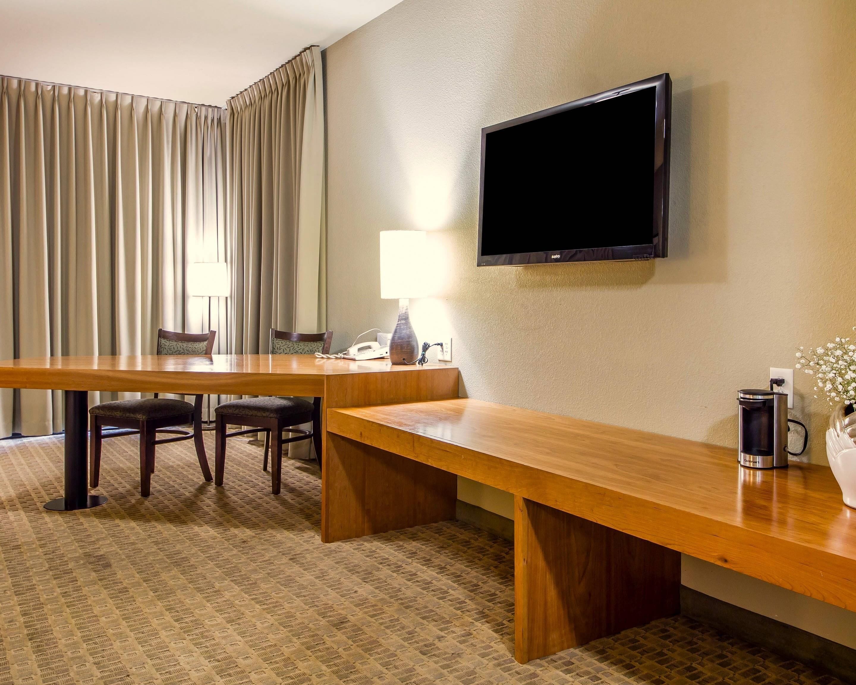 A hotel room with a wooden desk, a television mounted on the wall, a lamp, a coffee maker, and a window with curtains.