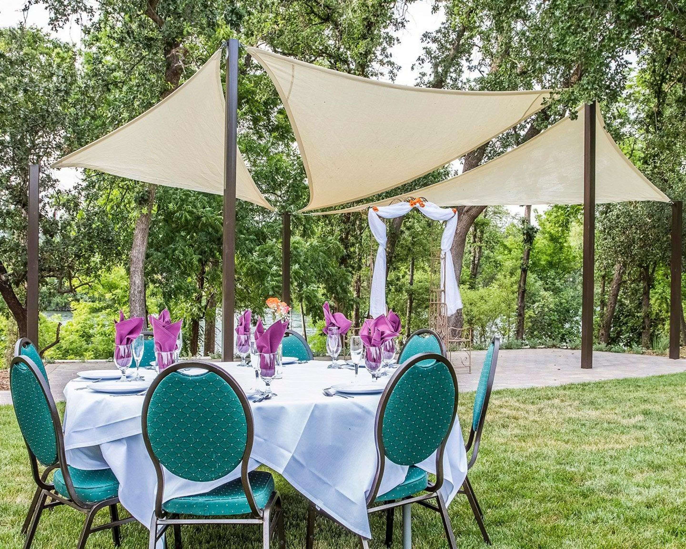 An outdoor dining area with round tables covered in white tablecloths, green upholstered chairs, and beige shade sails providing cover.