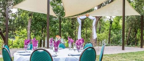 An outdoor dining area with round tables covered in white tablecloths, green upholstered chairs, and beige shade sails providing cover.