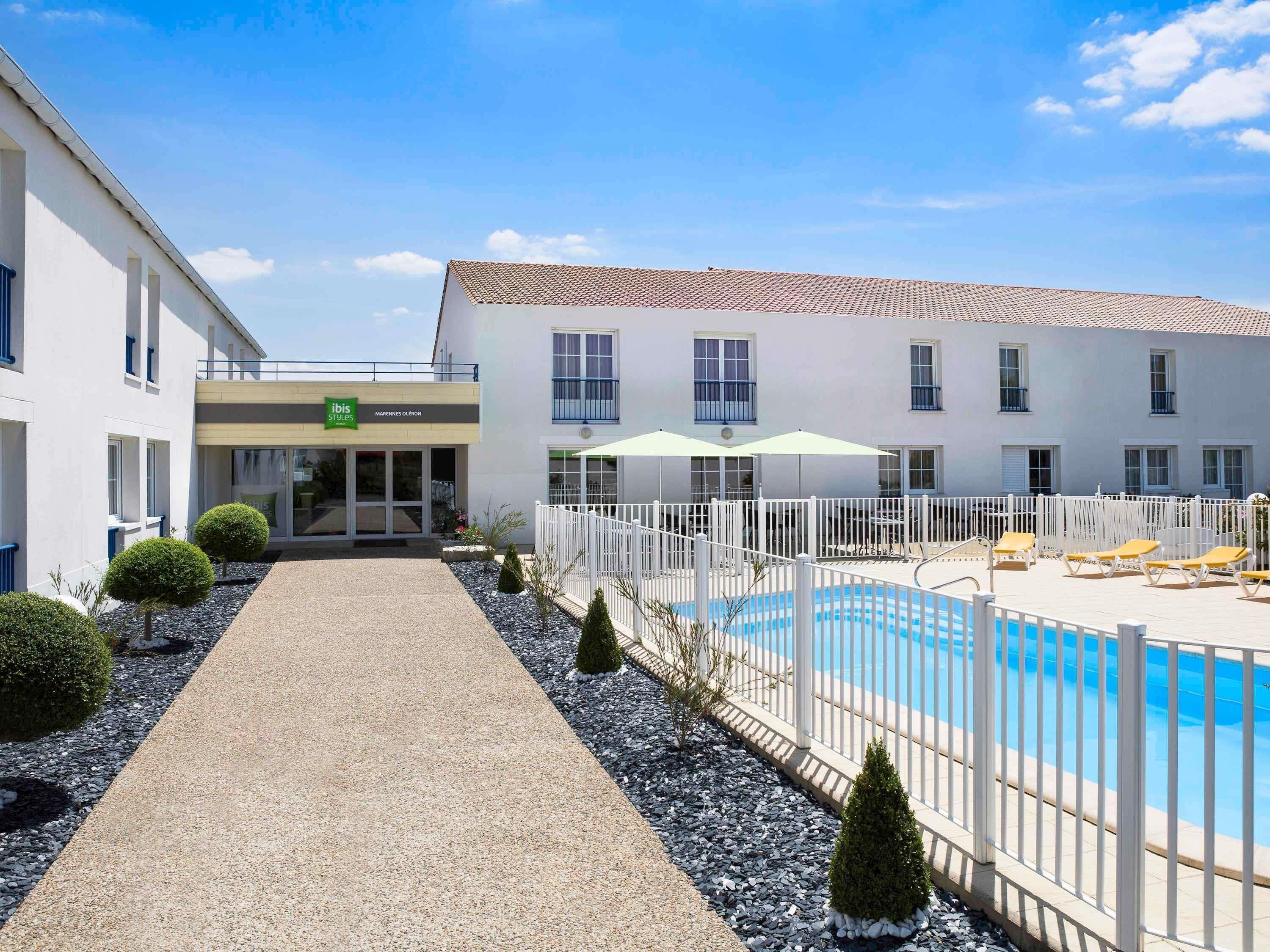 Un área de piscina con valla blanca, piscina y un edificio con un letrero que dice 'ibis Styles'.
