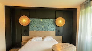 A hotel room with a large bed, two wall-mounted lamps, a round wooden table, and a patterned wall above the bed. - Hôtel ibis Styles Marennes Oleron (Marennes-Hiers-Brouage)