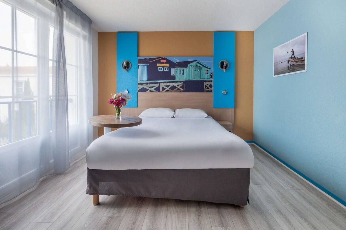 Ein Hotelzimmer mit einem Bett, einem runden Tisch mit Blumen in einer Vase, einem Gemälde an der Wand und einem Fenster mit Vorhängen.