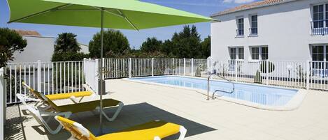 A poolside area with sun loungers, a green umbrella, and a white building with windows.