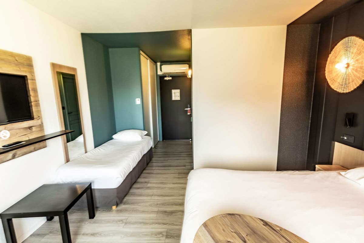 Une chambre d’hôtel avec deux lits simples, un petit bureau, un téléviseur fixé au mur et un couloir menant à une autre chambre.