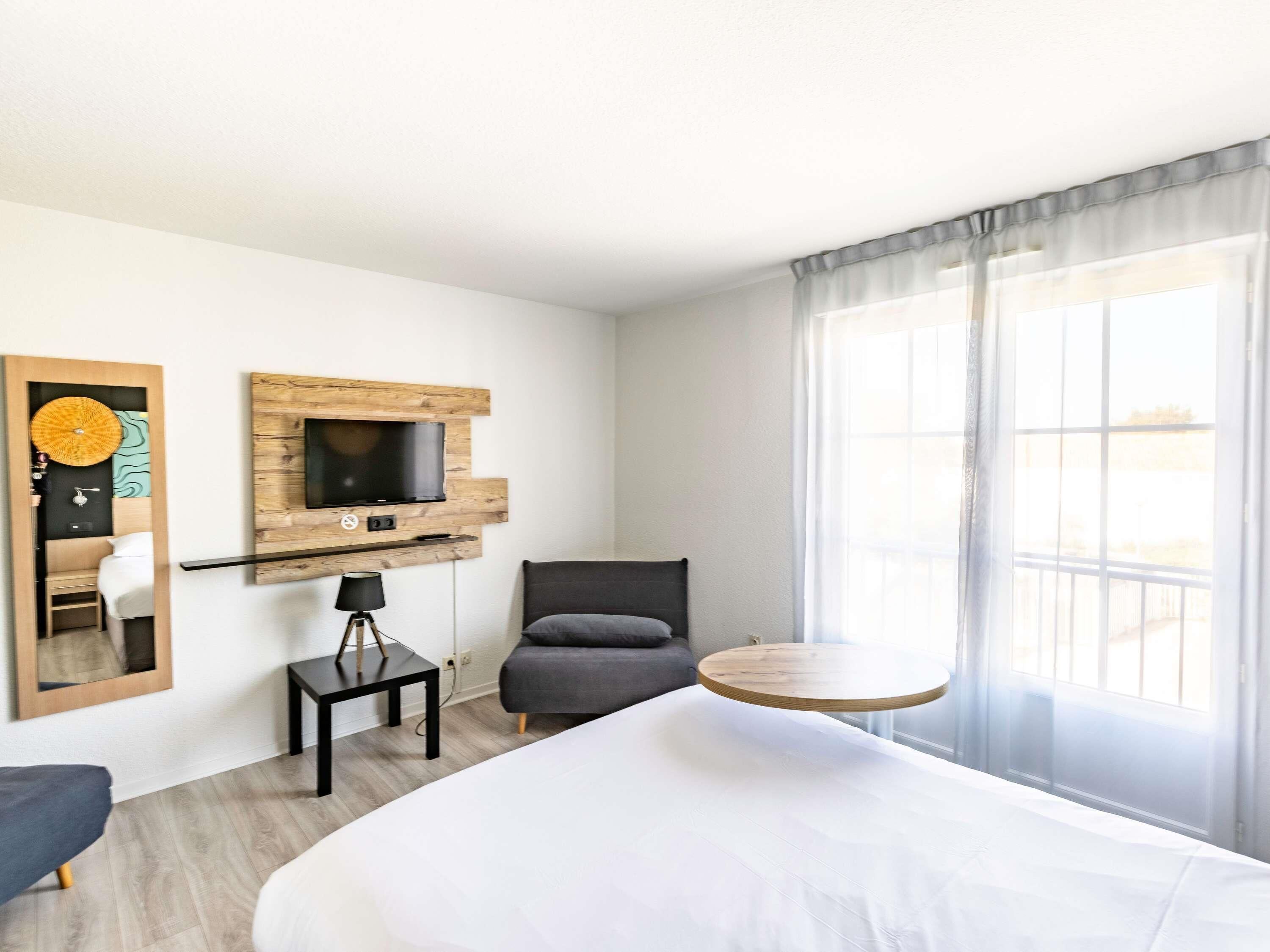 Una habitación de hotel con una cama, un televisor montado en un panel de madera, una silla, una mesita con una lámpara y una mesa redonda.