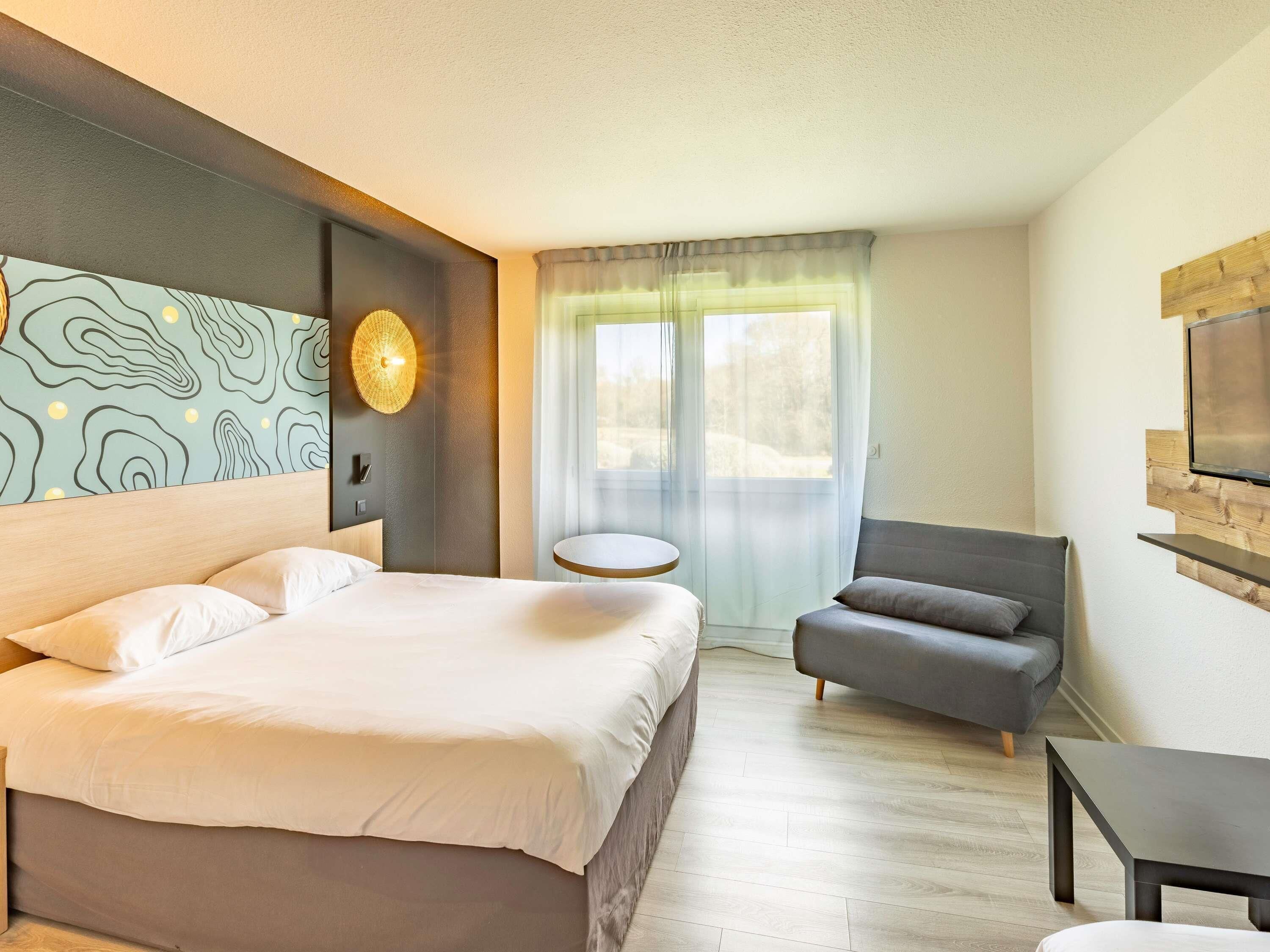 Una habitación de hotel con cama, una mesa redonda, un sillón gris, un televisor montado en una pared de madera y una ventana con cortinas de gasa.
