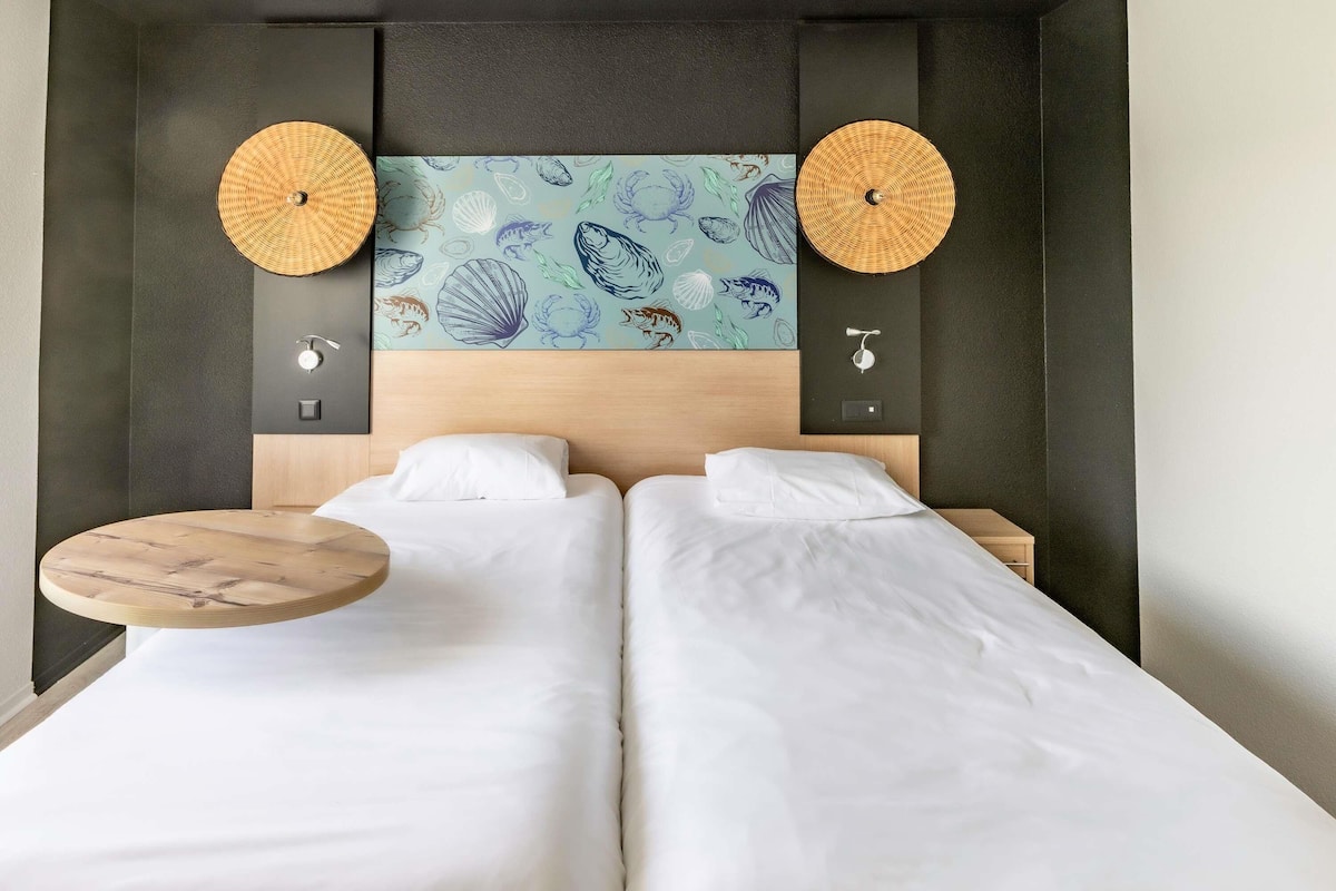 Cama doble con ropa de cama blanca, una mesa redonda de madera, dos lámparas de pared y una obra de arte decorativa con criaturas marinas.