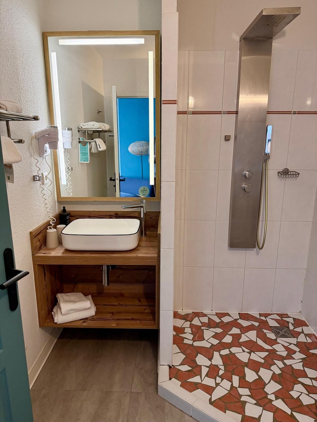 Un baño con tocador de madera, un lavabo blanco, un espejo, ducha con piso de azulejos y una puerta azul al fondo.