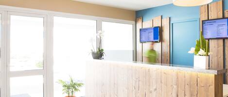 Zona de recepción de un hotel con un mostrador de madera, una pantalla digital que muestra tarifas de habitaciones y una planta en maceta.
