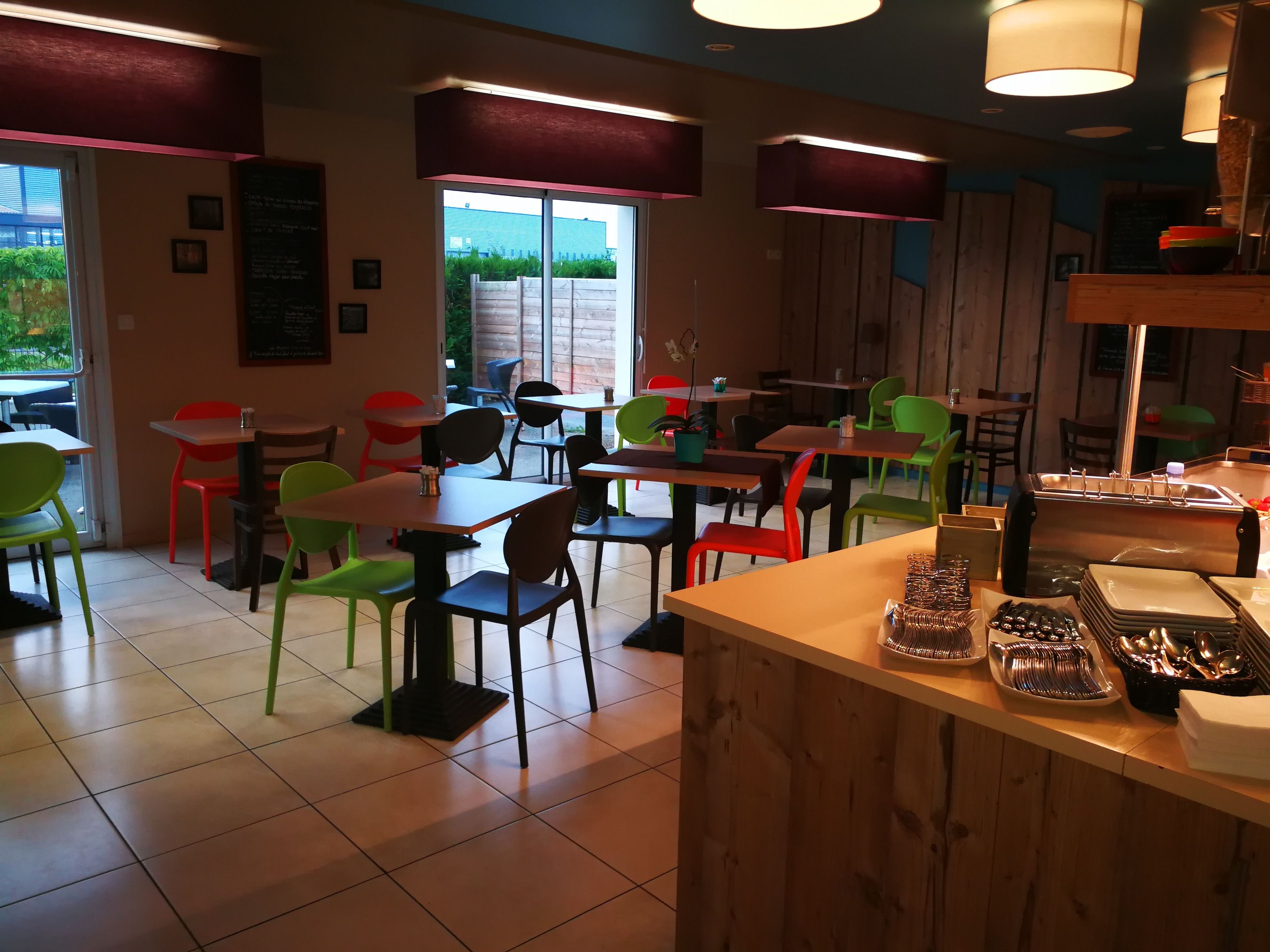 A restaurant interior with multiple tables and chairs, a counter with cutlery, and a view of greenery outside.