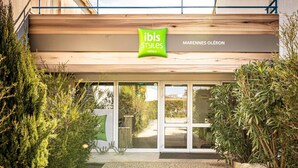 A hotel entrance with a wooden facade, a green sign for ibis styles hotels, and a gravel pathway leading to the entrance. - Hôtel ibis Styles Marennes Oleron (Marennes-Hiers-Brouage)