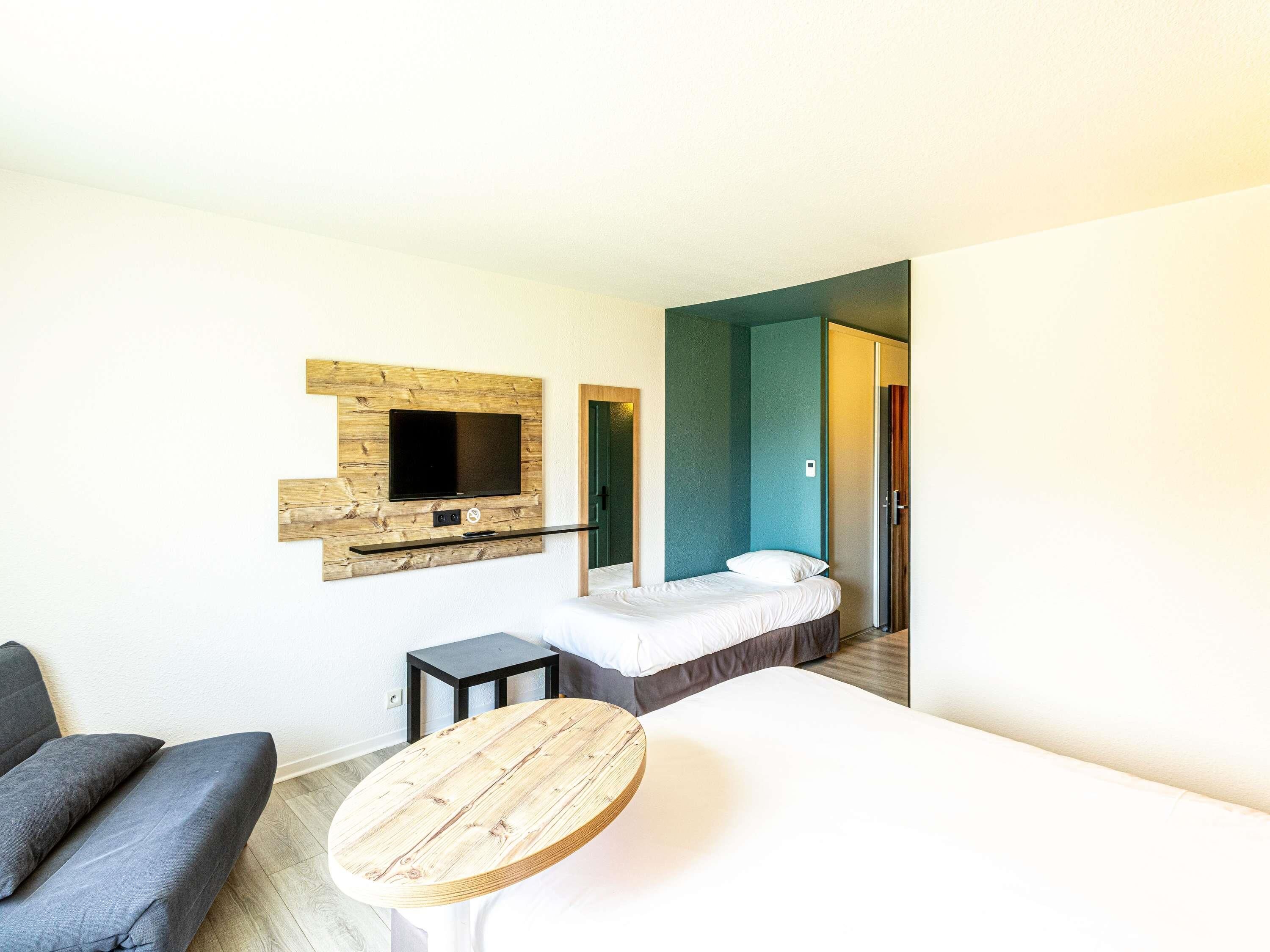 A hotel room with a bed, a TV mounted on a wooden panel, a small table, and a door leading to another room.