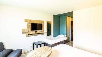 A hotel room with a bed, a TV mounted on a wooden panel, a small table, and a door leading to another room.