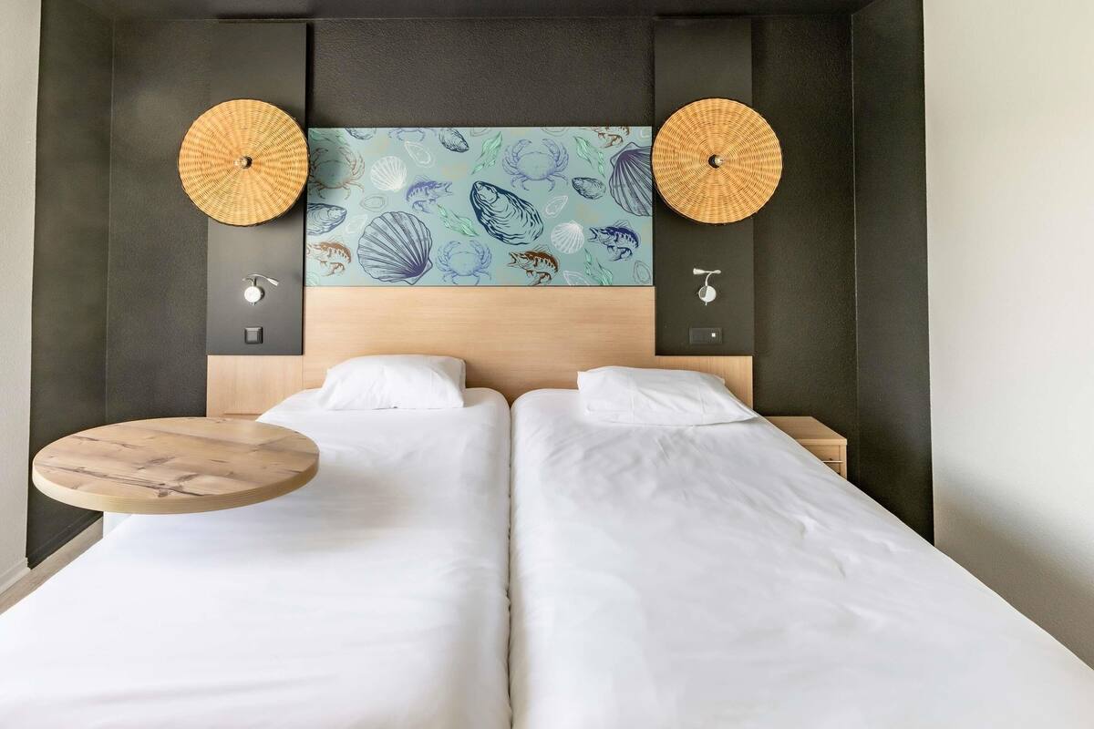 Un lit double avec du linge de lit blanc, une table ronde en bois, deux lampes fixées au mur et une décoration murale représentant des créatures marines.
