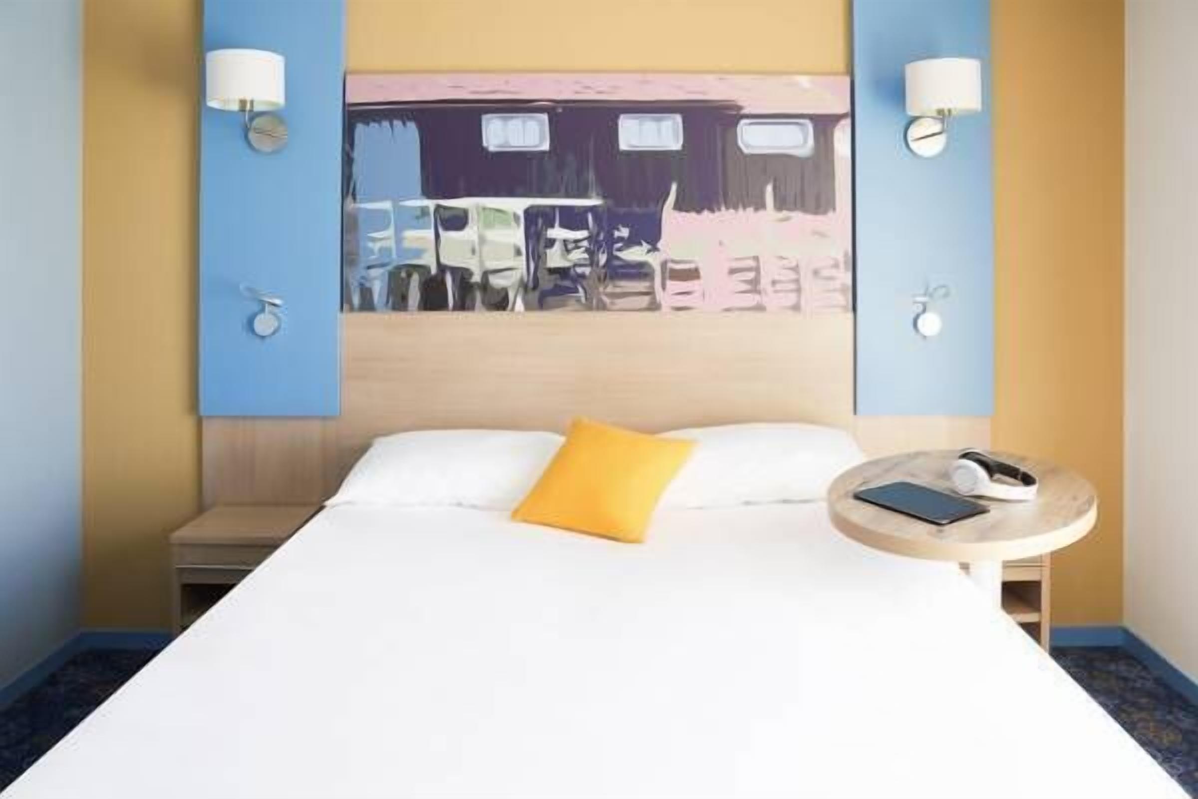 A hotel room with a bed, a yellow pillow, a bedside table with a phone and headphones, and a painting on the wall.