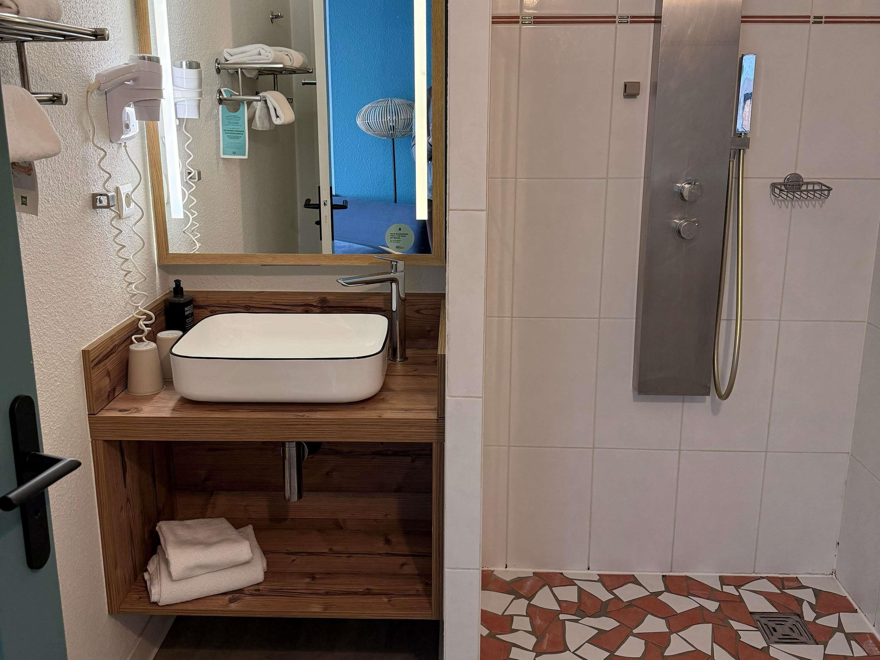 Une salle de bain avec un meuble-lavabo en bois, un lavabo blanc, un porte-serviettes, un miroir et une douche dotée d’une porte en verre.