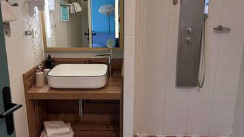 Une salle de bain avec un meuble-lavabo en bois, un lavabo blanc, un porte-serviettes, un miroir et une douche dotée d’une porte en verre.