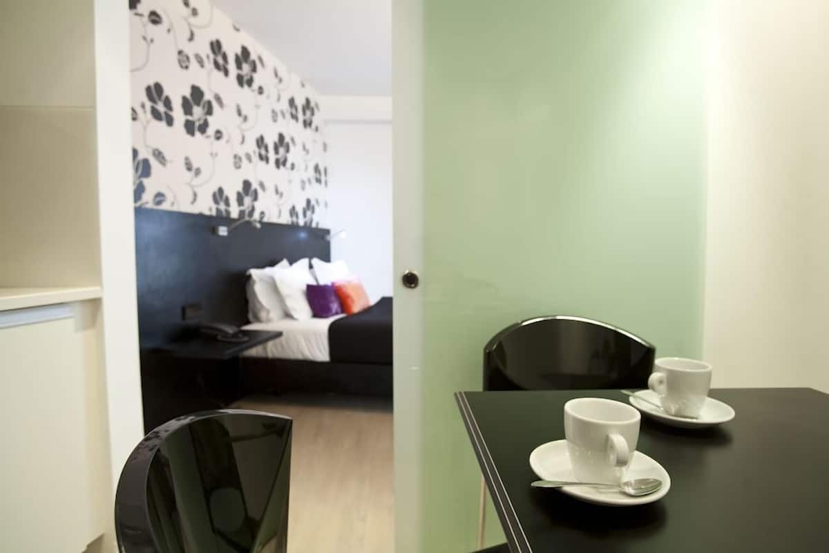 Ein Zimmer mit einem schwarzen Tisch, gedeckt mit zwei weißen Tassen und Untertellern, einem schwarzen Stuhl und einem Bett mit einem gemusterten Kopfteil.