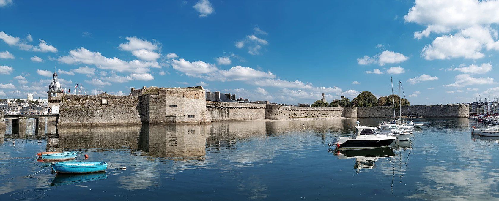 A historic stone wall with a moat, boats, and a clear sky.