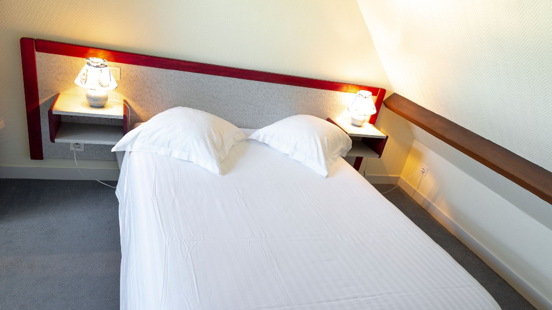 Una cama individual con ropa blanca, en una habitación con cabecera de madera y dos mesitas de noche con lámparas.
