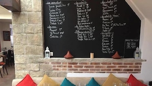 Un restaurant dont le menu est écrit à la craie sur un tableau noir, indiquant différents plats et leurs prix. Les tables sont dressées avec des verres à vin, des verres à eau et des couverts.