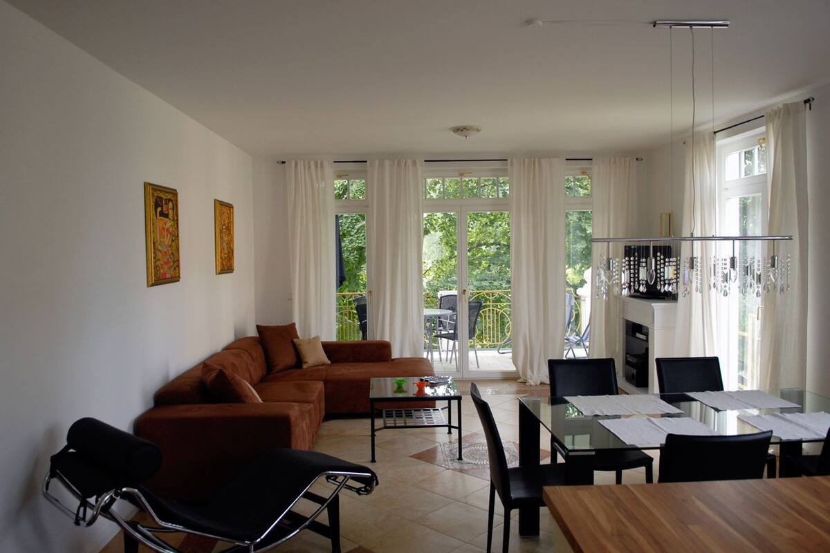 A modern living room with a brown sofa, black chairs, a glass dining table, and a balcony with white curtains.