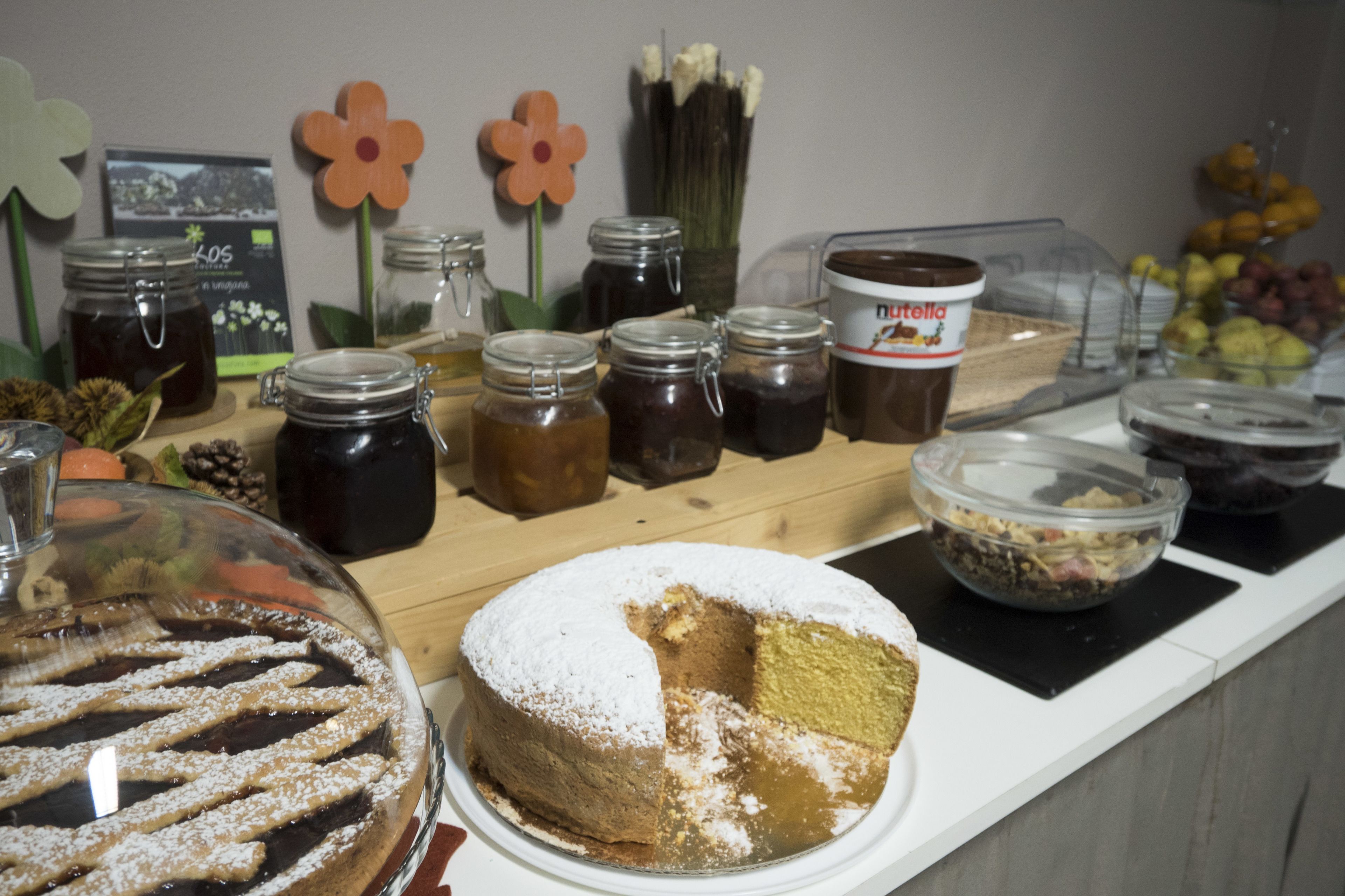 Una mesa de buffet con diversos alimentos, incluyendo un pastel con una porción faltante, frascos de conservas y un recipiente de Nutella.