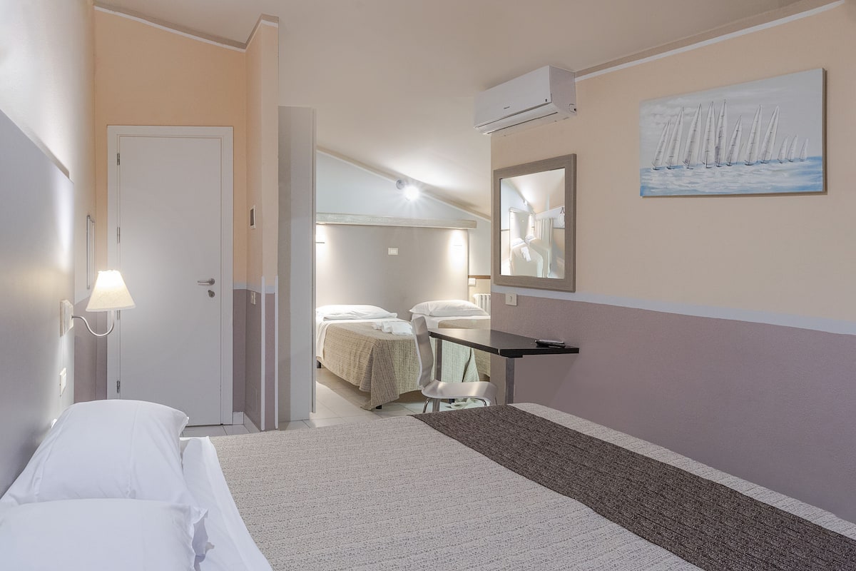 A hotel room with a bed, a desk, a chair, a lamp, an air conditioner, and a painting on the wall.