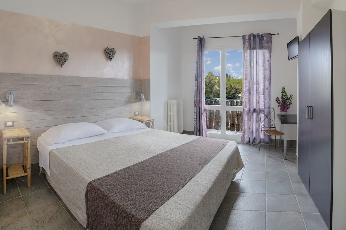 A bedroom with a bed, bedside tables, a nightstand, a dresser, a window with curtains, and a view of greenery outside.