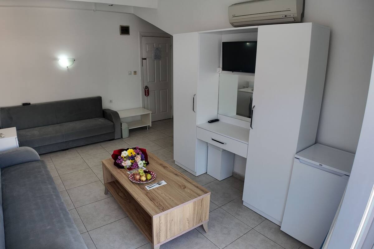Um espaço de estar compacto com sofá cinzento, mesa de centro de madeira com flores, guarda-roupa branco e uma TV montada sobre um móvel branco.