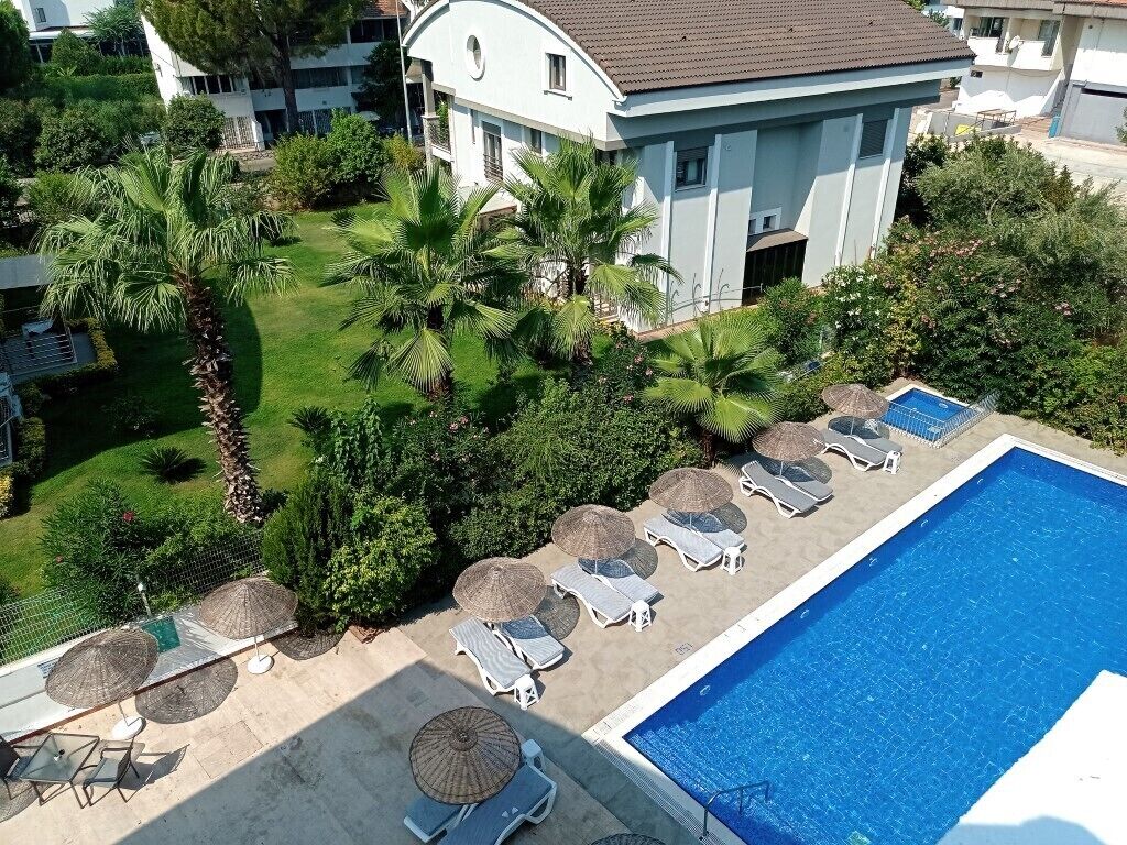A pool area with sun loungers and umbrellas, surrounded by greenery and a building with a curved roof.