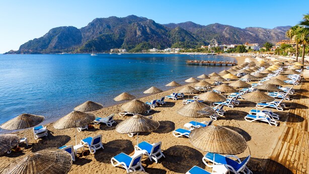 A beach with rows of sun loungers and thatched umbrellas, a clear blue sea, and mountains in the background. - Navy Hotel (Marmaris)