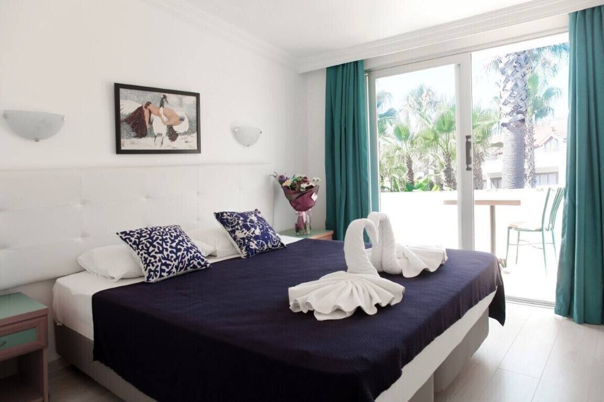 Quarto de hotel com cama, mesa de cabeceira, luminária na parede, quadro e vista para o exterior.