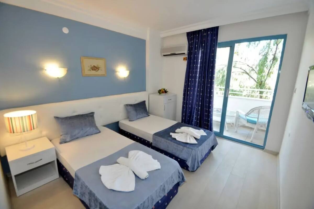 Quarto de hotel com duas camas, uma mesa de cabeceira, uma luminária e vista para o exterior através de uma porta de vidro.