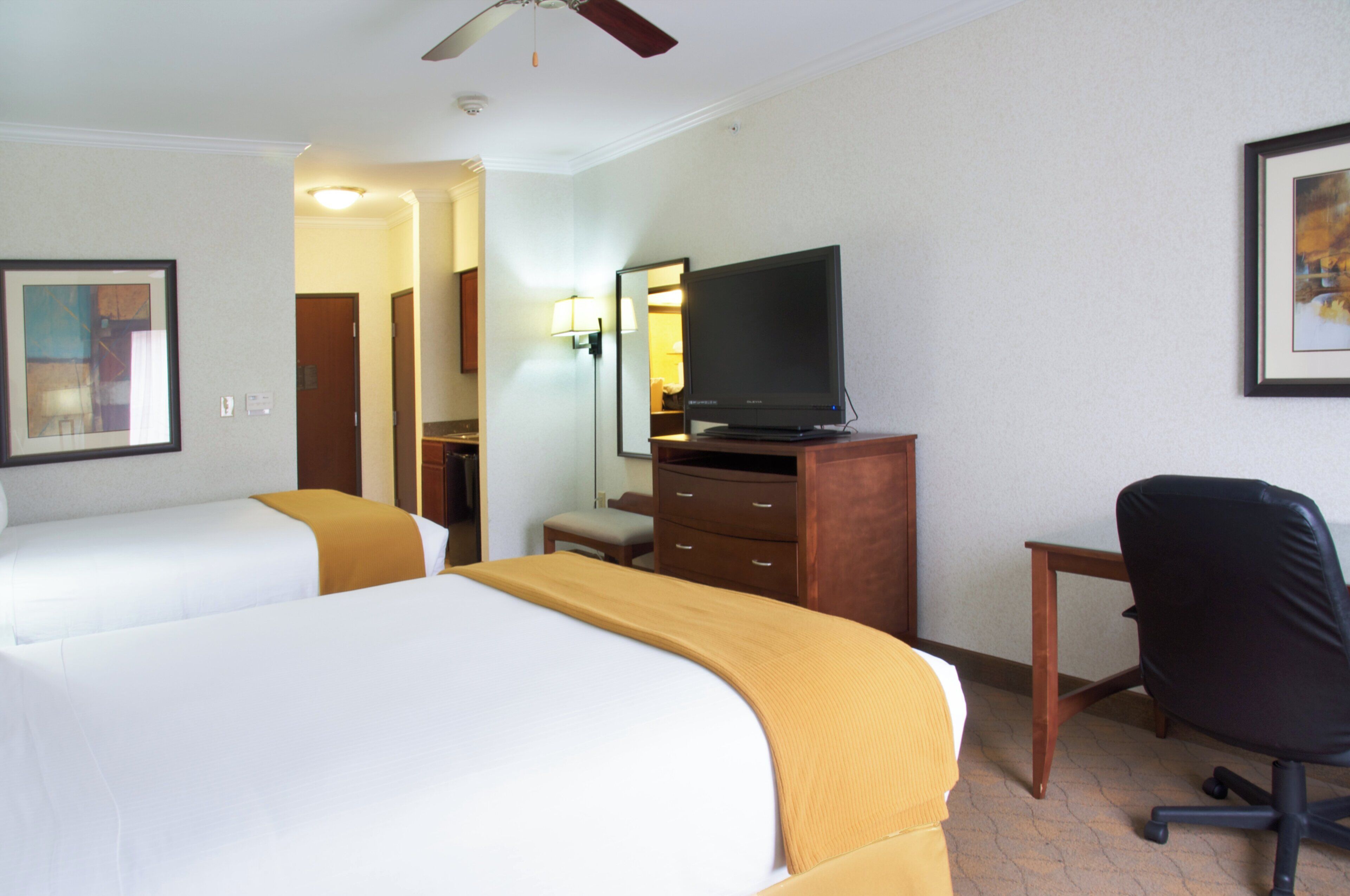 A hotel room with two beds, a television on a wooden dresser, a desk with a chair, and a ceiling fan.