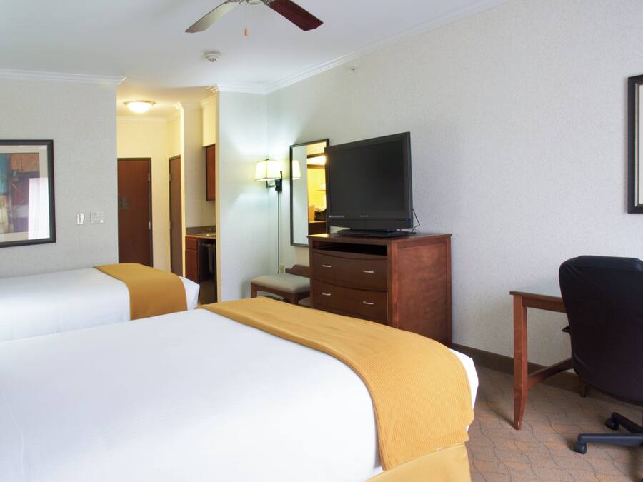A hotel room with two beds, a television on a wooden dresser, a desk with a chair, and a ceiling fan.