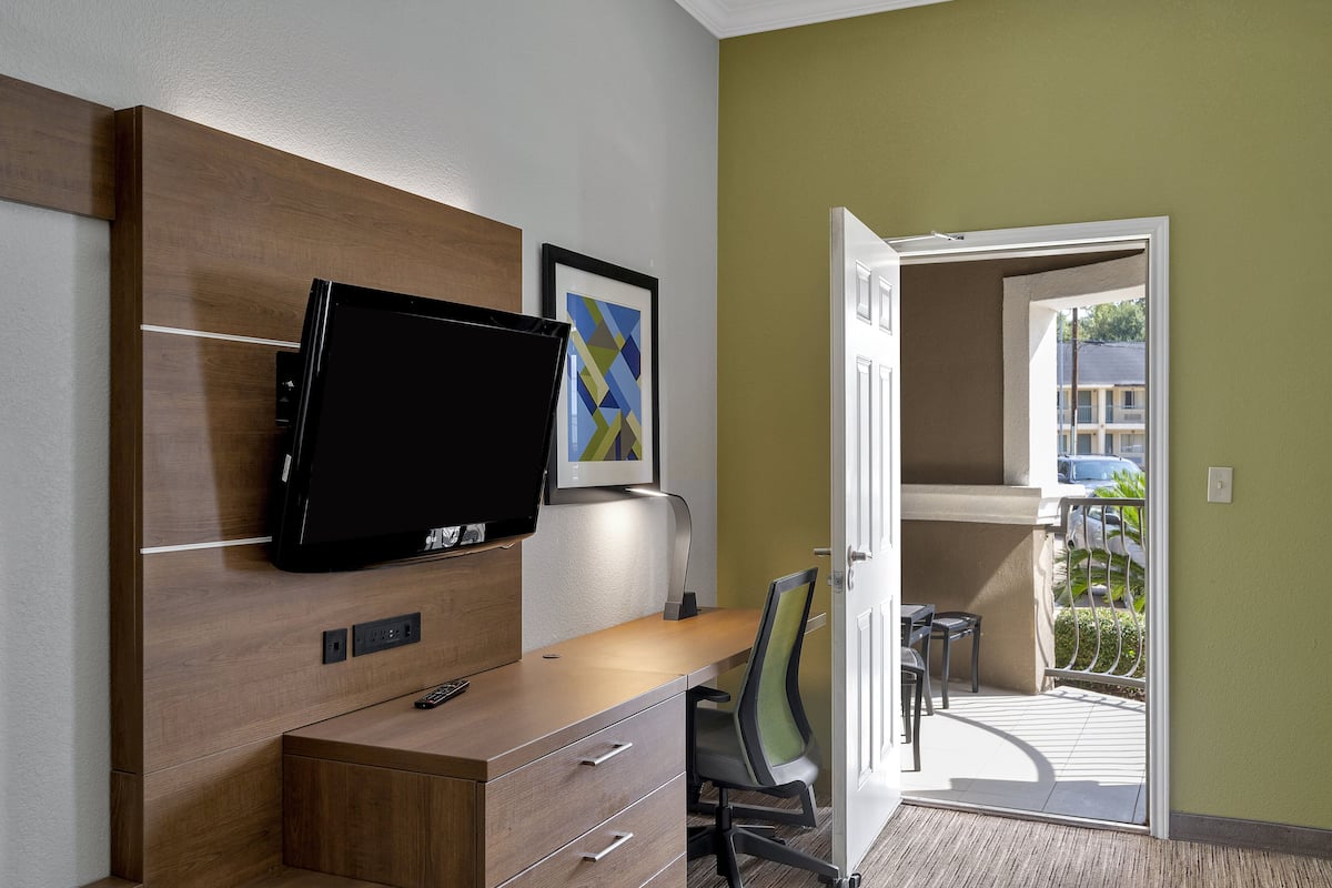 A hotel room with a wooden desk, a flat-screen TV, a framed artwork, and a view of the outdoor balcony.