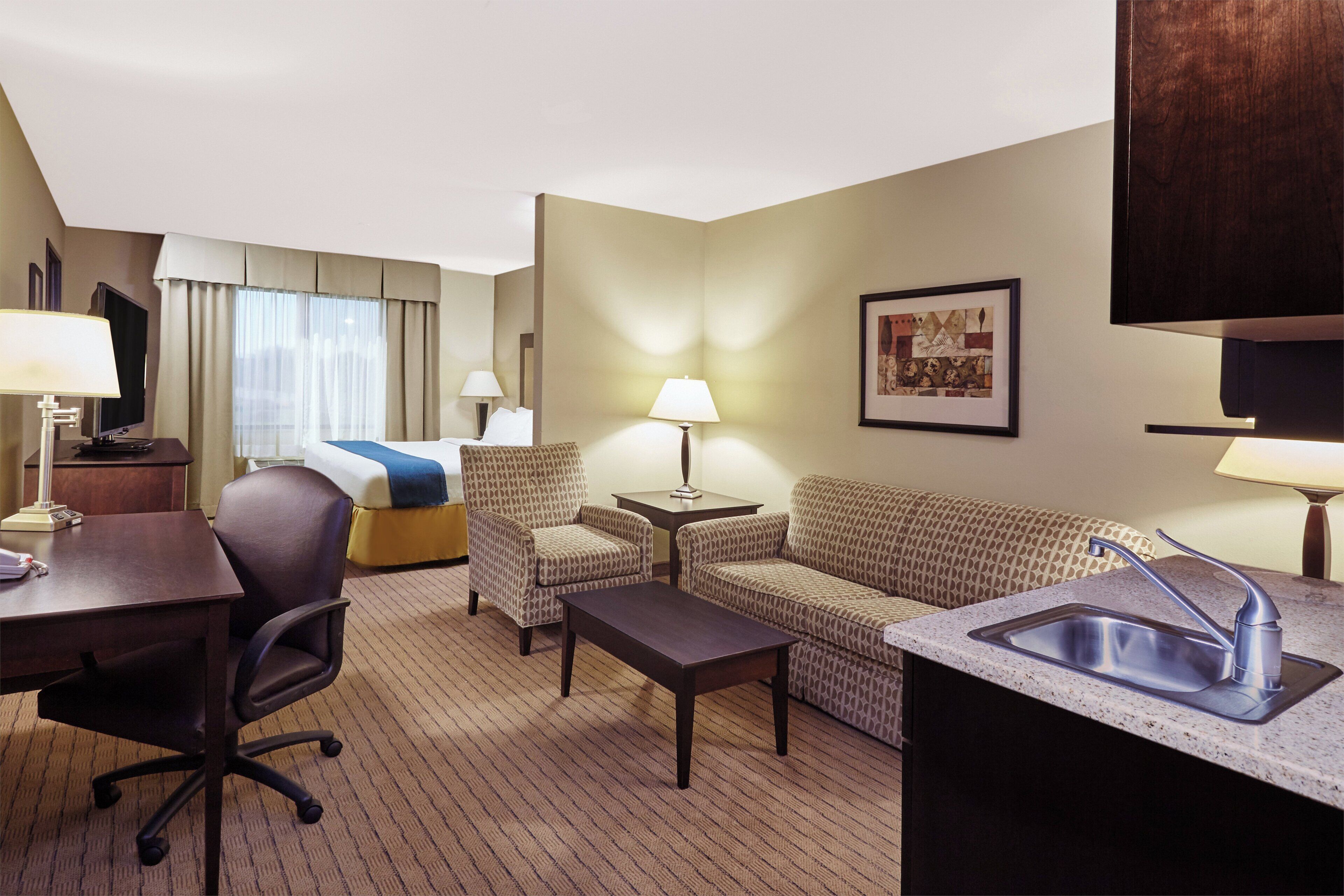 A hotel room with a bed, a desk, a chair, a sofa, a small table, a lamp, a picture on the wall, and a sink.