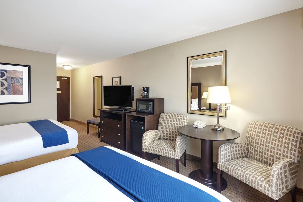 A hotel room with two beds, a desk with a TV, microwave, and telephone, two patterned armchairs, and a round table with a lamp.