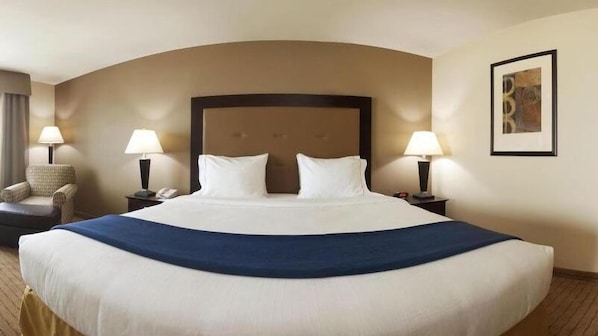 Una cama grande con sábanas blancas y una manta azul oscuro, flanqueada por dos mesitas de noche con lámparas, una silla y un cuadro en la pared.