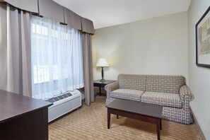 A hotel room with a sofa, a table, a lamp, a radiator, and curtains. - Holiday Inn Express Hotel & Suites Madison-Verona by IHG (Verona)