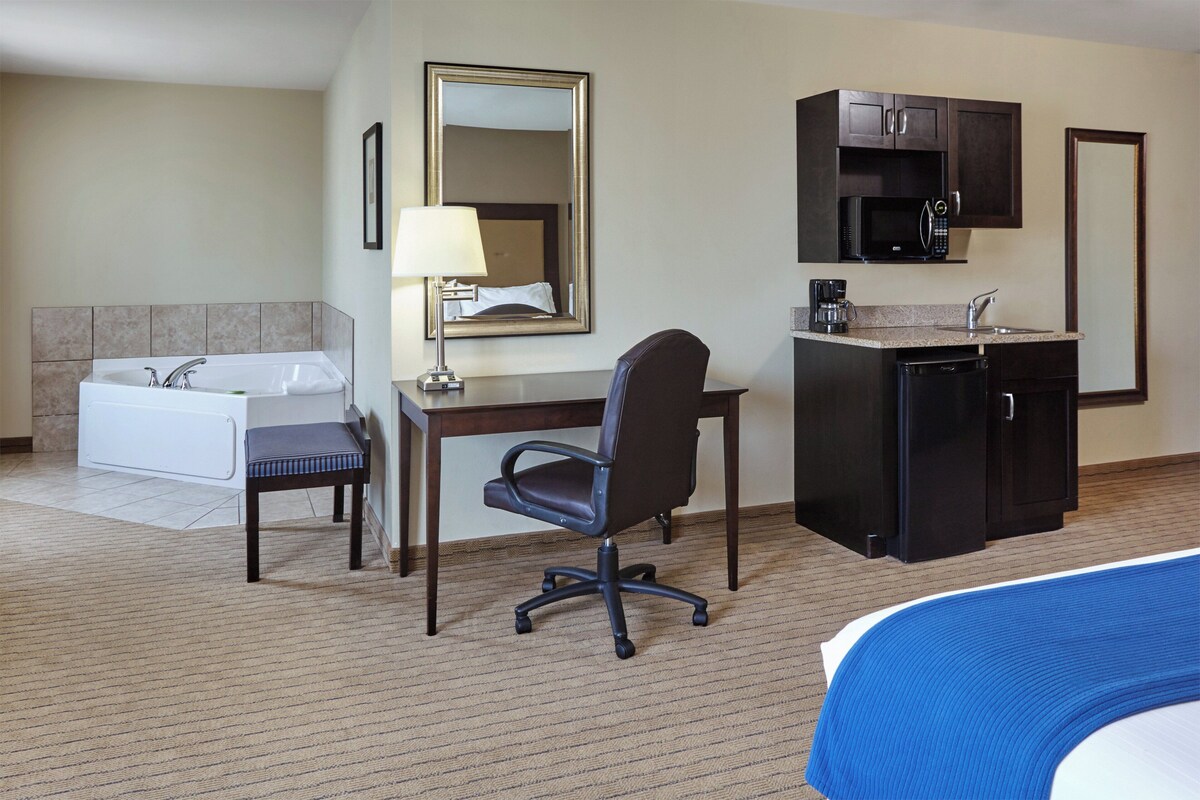 Une chambre d’hôtel comprenant un lit, un bureau, une chaise, une petite kitchenette, une salle de bain avec baignoire et un miroir.