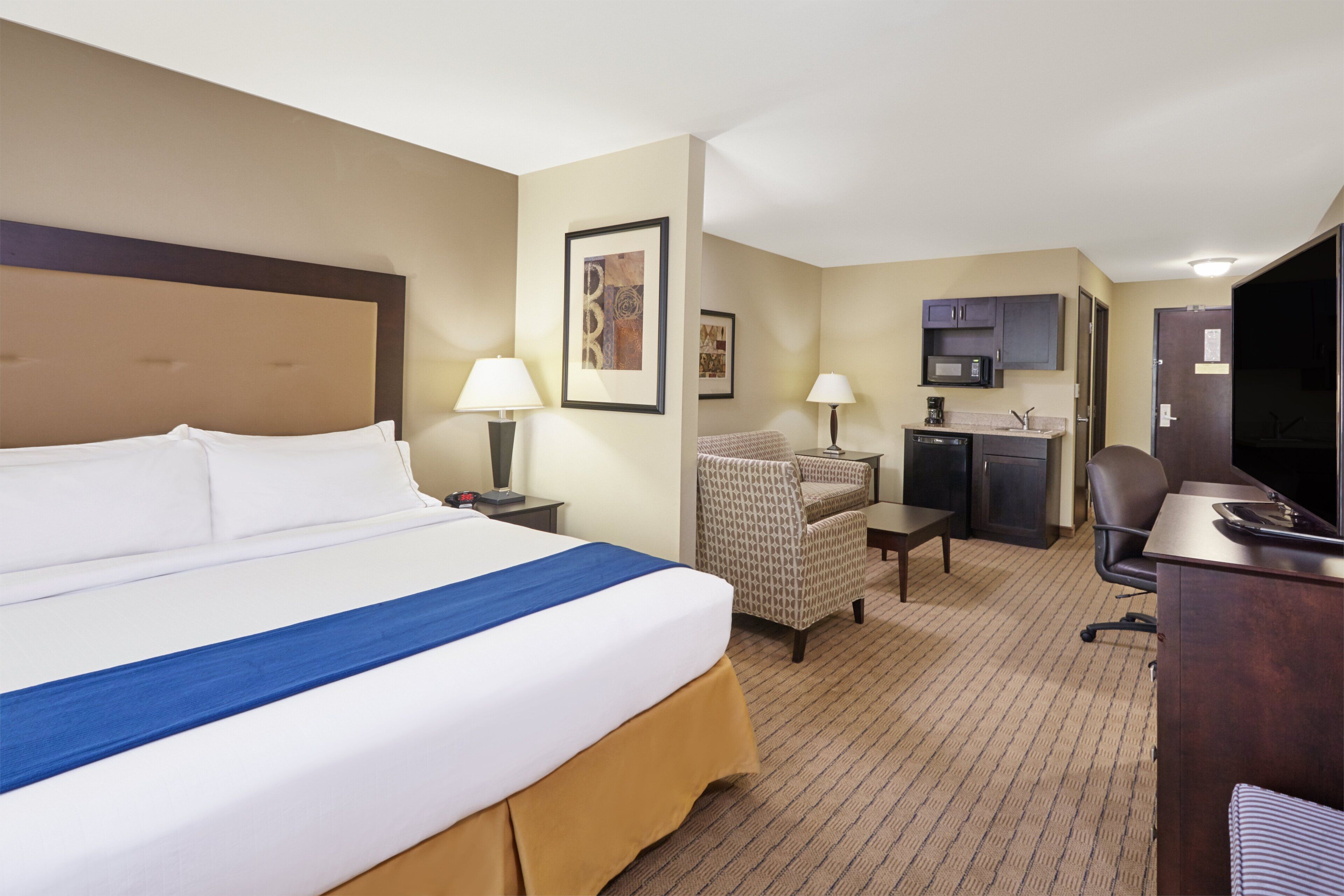 A hotel room with a large bed, a desk, a chair, a television, and a kitchenette.